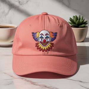 May include: A coral baseball cap with an embroidered clown face. The clown has a white face, red nose and mouth, and colourful hair and collar. The cap has a curved peak and a button on top.