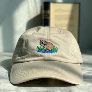 May include: A tan baseball cap with a curved brim. The cap features an embroidered raccoon design in brown, black, and white, sitting on blue water and green grass. The hat is made of a soft fabric.