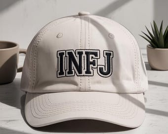 May include: A light beige baseball cap with the letters "INFJ" embroidered in black. The cap has a curved brim and a button on top. The background includes a coffee mug and a small potted plant.