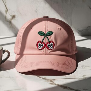 Puede incluir: Gorra de béisbol rosa claro con visera curva. La gorra presenta un diseño bordado de dos cerezas con caras de calaveras y hojas verdes. La gorra tiene un botón marrón en la parte superior y está hecha de un material suave.