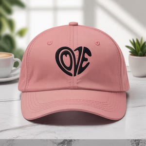 Può includere: Un cappellino da baseball rosa con visiera curva. Il cappellino presenta un design nero a forma di cuore con la parola "LOVE" in un carattere stilizzato. Il cappellino è realizzato in un materiale morbido e ha un bottone sulla parte superiore.