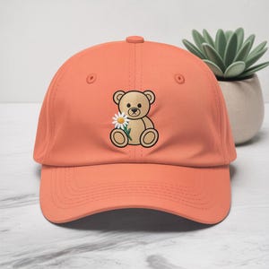 May include: Coral baseball cap with an embroidered teddy bear holding a daisy. The bear is light brown with black detailing. The cap has a curved peak and an adjustable strap at the back.