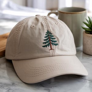Puede incluir: Gorra de béisbol beige con visera curva. La gorra presenta un diseño de árbol verde y marrón bordado en la parte delantera. La gorra está hecha de una tela suave y tiene un estilo clásico e informal.