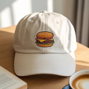 Pode incluir: Boné de beisebol bege com um bordado de hambúrguer de desenho animado. O boné tem uma aba curva e um fecho de tira de tecido. O design do hambúrguer inclui um pão de gergelim, queijo, alface e um hambúrguer.