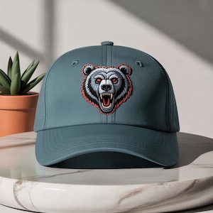 May include: A teal baseball cap with an embroidered bear head. The bear has red eyes, a snarling mouth, and white and grey fur outlined in orange. The cap has a curved peak and a button on top.