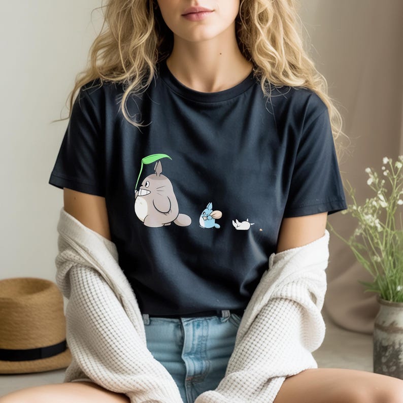 May include: A navy blue t-shirt featuring a cartoon illustration of three characters. The first is a gray, round creature holding a green leaf. The second is a small blue creature, and the third is a small white creature.