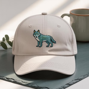 Puede incluir: Gorra de béisbol beige claro con un diseño de lobo bordado en color turquesa. El lobo tiene ojos amarillos y está de pie. La gorra tiene una visera curva y un botón en la parte superior. Una taza está en el fondo.