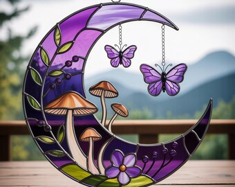 Peut inclure: Une lune en croissant en vitrail aux tons violets et mauves, ornée de champignons, d'une fleur et de deux papillons suspendus. La pièce décorative a un cadre en métal et un ruban violet pour la suspendre. L'arrière-plan montre une scène extérieure floue.