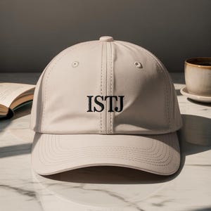 May include: A beige baseball cap with the embroidered letters "ISTJ" in black. The cap has a curved brim and a button on top. The background includes a book and a cup of coffee.