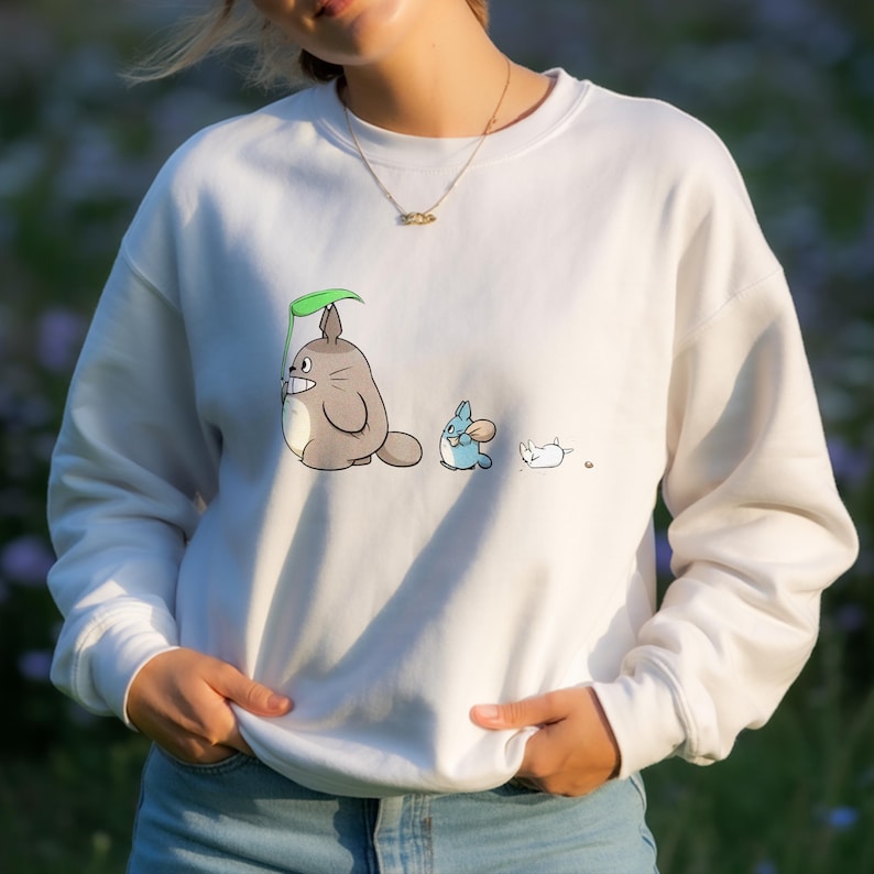 May include: White sweatshirt featuring a cartoon illustration of three characters. The first is a gray creature holding a green leaf, followed by a blue creature, and a white creature lying down. The sweatshirt has long sleeves and a crew neck.