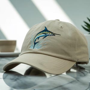 May include: A tan baseball cap featuring an embroidered marlin leaping from the water. The marlin is blue, yellow, and black, with light blue water details. The cap has a curved brim and a classic six-panel design.