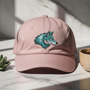 Pode incluir: Boné de beisebol rosa claro com um desenho de lobo bordado. O lobo é retratado em perfil com detalhes turquesa e brancos. O boné tem uma aba curva e uma alça ajustável.