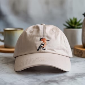 Puede incluir: Gorra de béisbol beige con visera curva. La gorra presenta un diseño de pájaro bordado en naranja, negro y blanco en la parte delantera. La gorra está hecha de una tela suave y tiene un diseño clásico de seis paneles.