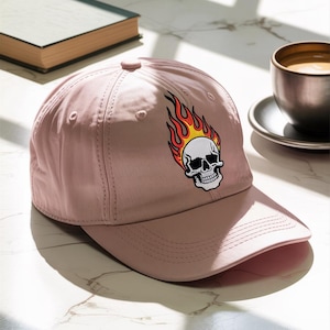 May include: A dusty rose baseball cap featuring an embroidered skull with flames. The skull is white with black details, and the flames are red and yellow. The cap has a curved brim and a button on top.