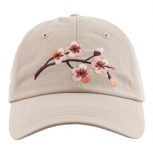 May include: A tan baseball cap featuring an embroidered cherry blossom branch with pink and peach flowers and brown stems. The cap has a curved brim and a classic six-panel design.
