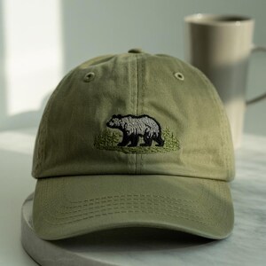May include: An olive green baseball cap with a curved peak. The cap has an embroidered design of a bear in grey and black, standing in front of green trees. The cap is made of a soft fabric and has a classic, casual style.