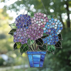 May include: Stained glass panel featuring a bouquet of hydrangeas in shades of purple, blue, and pink. The flowers are in a blue pot with green leaves. The panel is designed to hang and is suspended by a chain.