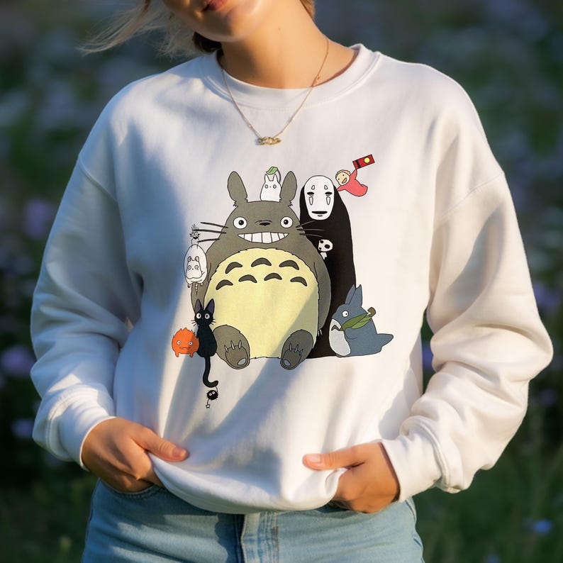 May include: White sweatshirt featuring a colorful graphic of animated characters. The design includes a large gray creature with a yellow belly, a black figure, and other smaller characters. The sweatshirt has long sleeves and a crew neck.