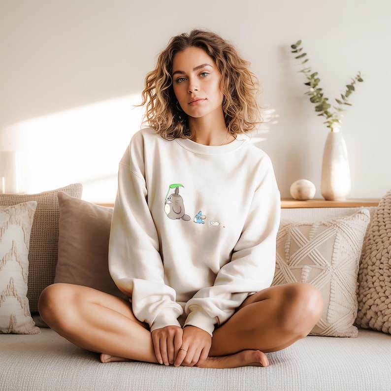 May include: Cream-colored sweatshirt featuring a cartoon illustration of a gray creature with a green leaf, a blue creature, and a white creature. The sweatshirt is a casual, long-sleeved garment.