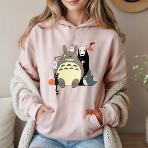 May include: A light pink hoodie featuring a colorful graphic of animated characters. The graphic includes a large gray creature with a wide smile, a black figure, and other smaller characters. The hoodie has a kangaroo pocket and a drawstring hood.