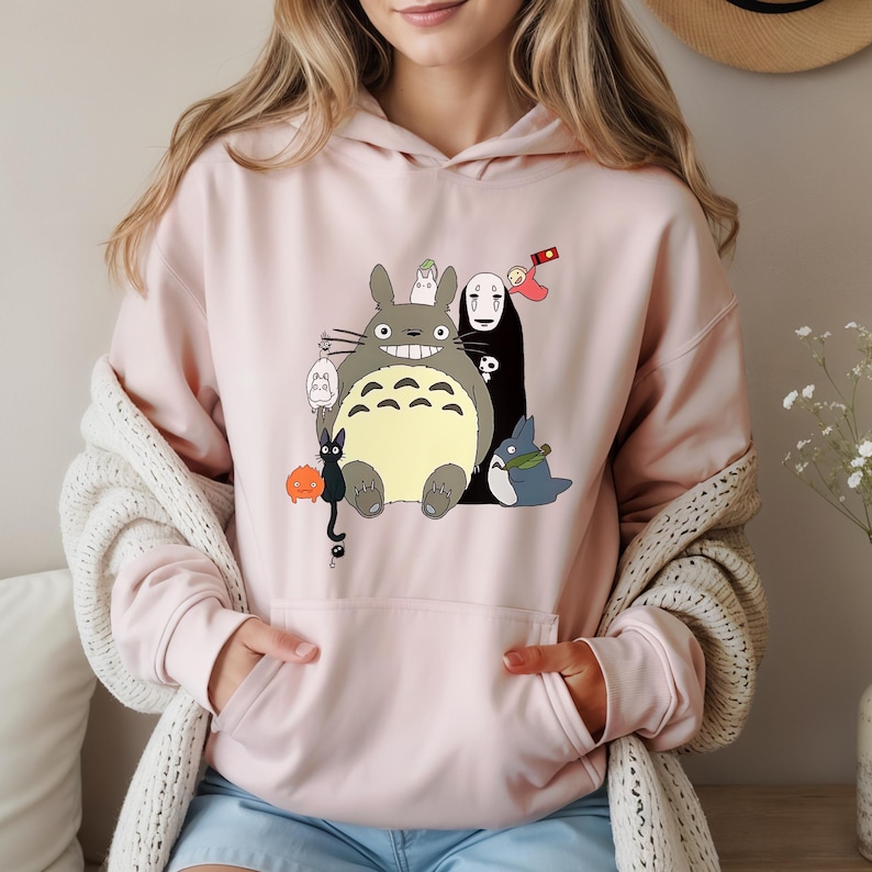 May include: A light pink hoodie featuring a colorful graphic of animated characters. The graphic includes a large gray creature with a wide smile, a black figure, and other smaller characters. The hoodie has a kangaroo pocket and a drawstring hood.