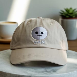 Puede incluir: Gorra de béisbol beige con visera curva. La gorra presenta una cara bordada en blanco con un ojo negro y una X, y la palabra "VAN" bordada debajo. La gorra está sobre una superficie de mármol.