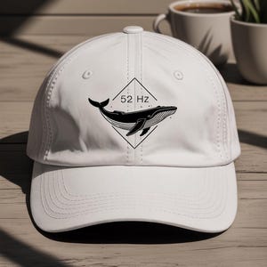 May include: White baseball cap with a black and white whale design inside a diamond, and the text "52 Hz". The cap has a curved peak and a button on top. A casual accessory.