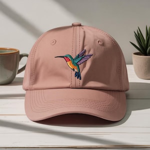 May include: A dusty rose baseball cap featuring an embroidered hummingbird. The hummingbird has a red throat, green head, orange chest, and blue wings. The cap has a curved brim and a button on top.
