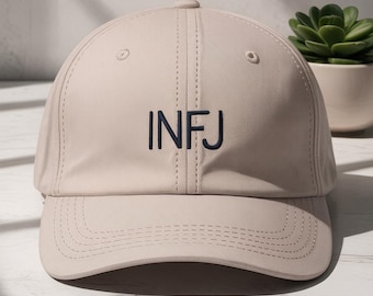 May include: A beige baseball cap with the embroidered letters "INFJ" in navy blue. The cap has a curved brim and a button on top. The background includes a potted succulent and a marble surface.
