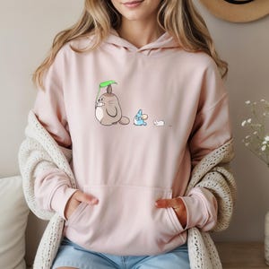 May include: A light pink hoodie with a kangaroo pocket and a drawstring hood. The front features a cartoon illustration of three characters: a large, gray creature holding a leaf, a blue creature, and a small white creature.