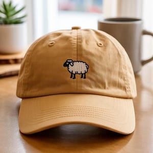 Könnte beinhalten: Beige Baseball Cap mit gebogenem Schirm. Die Kappe hat ein kleines, gesticktes Schafdesign in Schwarz und Weiß auf der Vorderseite. Die Kappe ist aus weichem Stoff und hat ein klassisches Sechs-Panel-Design.