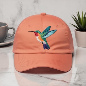 May include: A coral-colored baseball cap featuring an embroidered hummingbird. The hummingbird has a long black beak, a red throat, and a colorful body with blue, green, orange, and white details. The cap has a curved brim and a button on top.