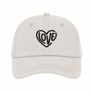 May include: A light beige baseball cap with a curved brim. The cap features a black heart-shaped design with the word "LOVE" in a stylized font. The cap has a button on top and stitched details.
