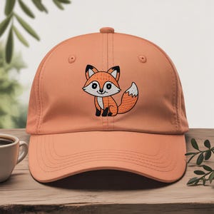 Op de afbeelding: Perzik kleurige baseball cap met een geborduurde cartoon vos. De vos heeft oranje vacht, een witte borst en een pluizige witgepunte staart. De cap heeft een gebogen klep en een knoop aan de bovenkant.