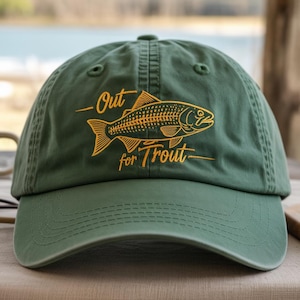 May include: Green baseball cap with the text "Out for Trout" and a gold fish illustration. The cap is made of a soft material and has a curved brim. The hat is sitting on a wooden surface.