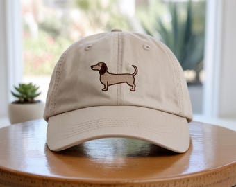 Puede incluir: Gorra de béisbol beige con un diseño de perro salchicha bordado en marrón. La gorra tiene una visera curva y un cierre de correa de tela. El perro está representado de pie con la cola levantada. La gorra está sobre una mesa de madera.