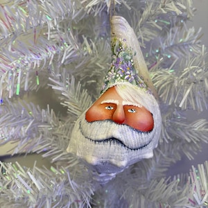 May include: A hand-painted Christmas ornament shaped like Santa Claus. The ornament features a white beard and eyebrows, rosy cheeks, and blue eyes. The top of the ornament is decorated with purple and white flowers. It hangs from a thin, natural-colored string.