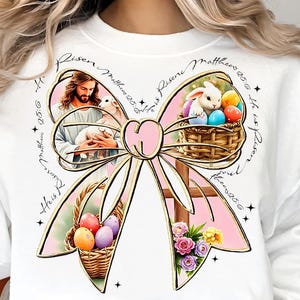 May include: White sweatshirt with a pink bow design. The bow has images of Jesus, a lamb, a basket of Easter eggs, and a cross. The text "He is Risen Matthew 20:6" is repeated around the bow.