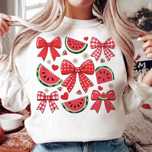 Pode incluir: Camisola branca com um padr&atilde;o de melancia e la&ccedil;o vermelho e branco. O design apresenta la&ccedil;os de bolinhas vermelhas e brancas, la&ccedil;os xadrez vermelhos e brancos e fatias de melancia.
