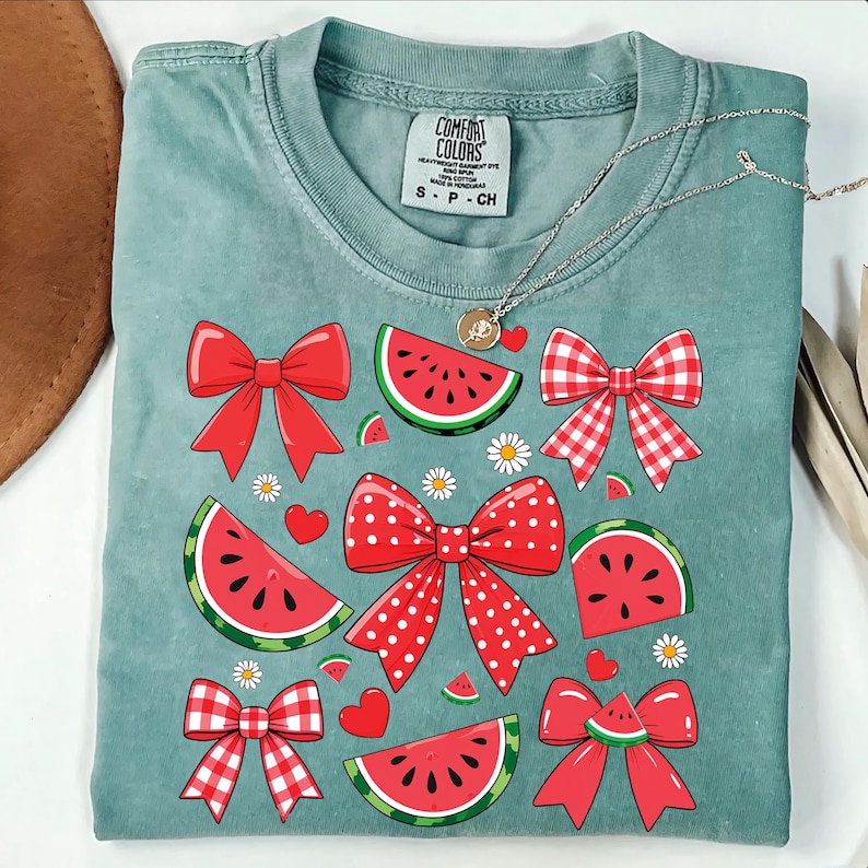Pode incluir: Uma camiseta azul turquesa com um design gr&aacute;fico de la&ccedil;os vermelhos, brancos e rosa e fatias de melancia. A camiseta tem o texto "COMFORT COLORS" na etiqueta.