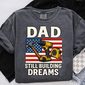 Könnte beinhalten: Dunkelgraues Sweatshirt mit dem Wort "DAD" über einer amerikanischen Flagge. Darunter steht "STILL BUILDING DREAMS". Die Flagge zeigt Hammer, Bohrmaschine und Maßband.