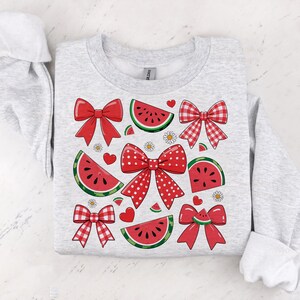Pode incluir: Uma camisola cinzenta com um design de melancia e la&ccedil;os. A melancia &eacute; vermelha e verde com sementes pretas. Os la&ccedil;os s&atilde;o vermelhos e brancos com bolinhas brancas.