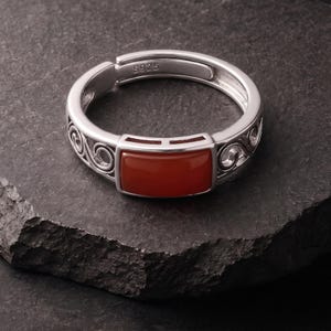 Könnte beinhalten: Ein silberner Ring mit einem rechteckigen, glatten, tief orangefarbenen Stein in der Mitte. Die Ringschiene hat ein offenes, verstellbares Design und zeigt auf den Seiten komplizierte, geschwungene Verzierungen. Innen ist S925 eingeprägt.