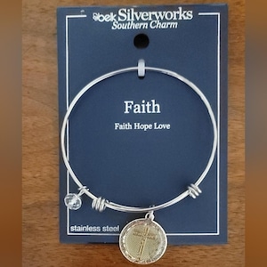 May include: A silver-tone stainless steel bracelet with a round charm featuring a cross design. The bracelet has the word "Faith" and "Faith Hope Love" printed on the dark blue card. A clear crystal bead hangs from the bracelet.