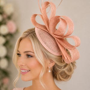 May include: A peach-coloured fascinator hat featuring a circular base and large, structured loops made of a woven, textured fabric. The headpiece is positioned on the side of the head, showcasing a symmetrical arrangement of stiffened, curved ribbons.