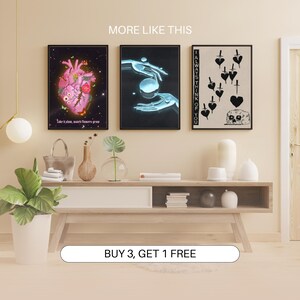 May include: Three framed art prints with different designs. The first print features a pink anatomical heart with flowers and butterflies. The second print shows a pair of hands holding a glowing orb. The third print is black and white and features a skull with a heart and daggers. The text "I ALWAYS THINK OF YOU" is on the right side of the print. The prints are displayed on a wooden shelf with a wooden table below.