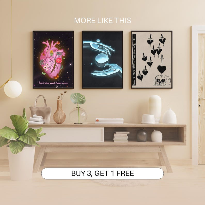 May include: Three framed art prints with different designs. The first print features a pink anatomical heart with flowers and butterflies. The second print shows a pair of hands holding a glowing orb. The third print is black and white and features a skull with a heart and daggers. The text "I ALWAYS THINK OF YOU" is on the right side of the print. The prints are displayed on a wooden shelf with a wooden table below.