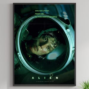May include: A framed movie poster for the film "Alien" featuring a close-up of a person in a spacesuit helmet. The poster has a dark green and black color scheme with the film's title and tagline text.