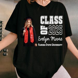 May include: Black t-shirt featuring a photo of a graduate wearing a cap and red sash. The shirt has the text "CLASS 2025 Evelyn Moore FLORIDA STATE UNIVERSITY" in white block letters. The graduate is smiling.