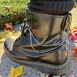 May include: Black leather combat boot with silver chain and star charm detail. The boot features a lace-up design and a yellow stitched sole. A black knit sock is visible at the top of the boot. Autumn leaves are scattered around the boot.